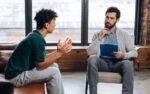Male bearded psychoanalyst holding clipboard and listening teenage boy in the office